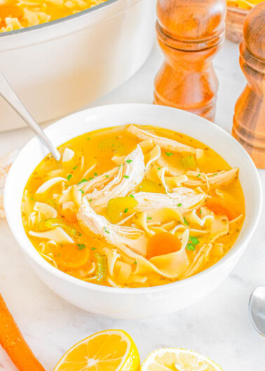 A bowl of chicken noodle soup with shredded chicken, noodles, and carrots, garnished with herbs, sits on a white surface next to a pot, two wooden pepper grinders, and halved lemons.