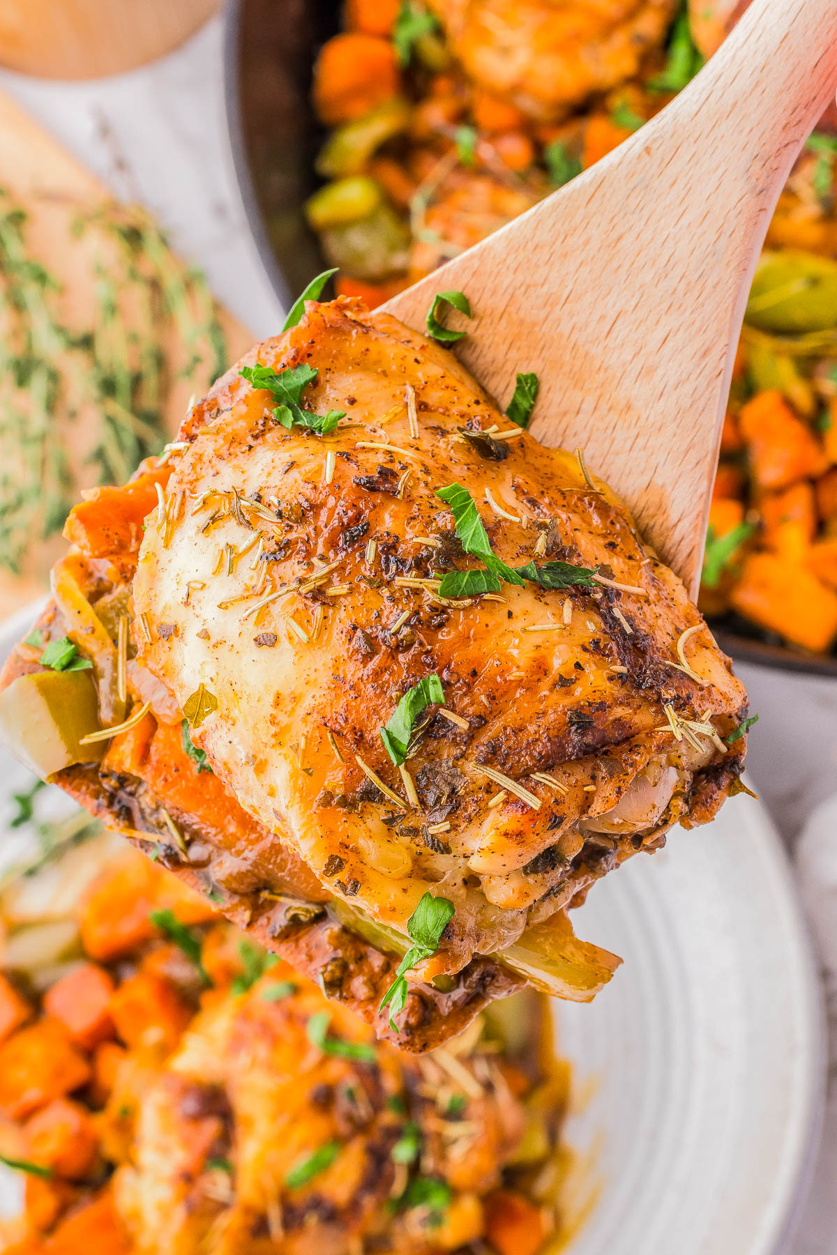 A wooden spoon holding a seasoned cooked chicken thigh garnished with herbs. In the background are more pieces of cooked chicken and vegetables on a plate.