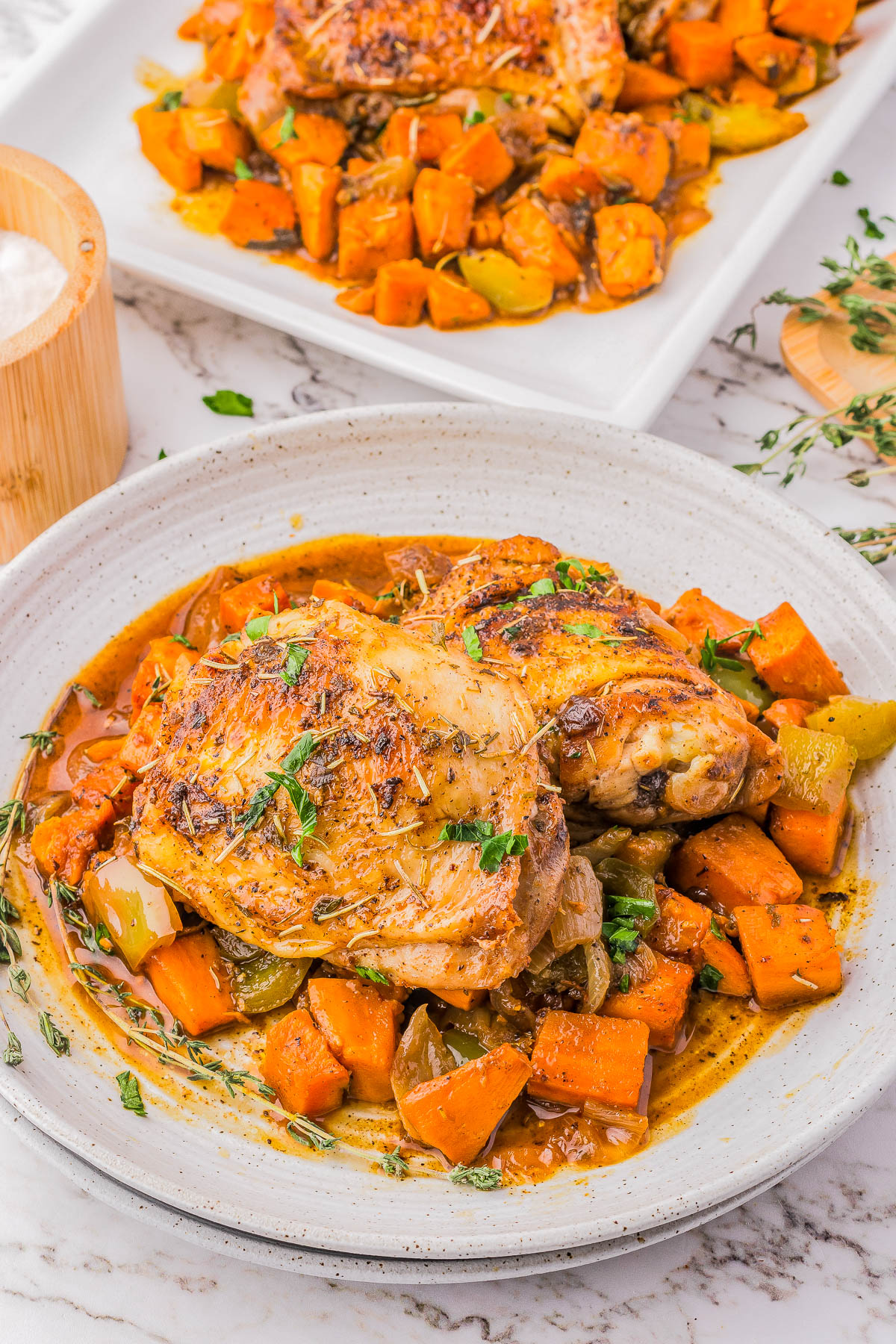 A white plate with roasted chicken pieces, garnished with herbs and served with chunks of sweet potatoes and vegetables in a savory sauce.