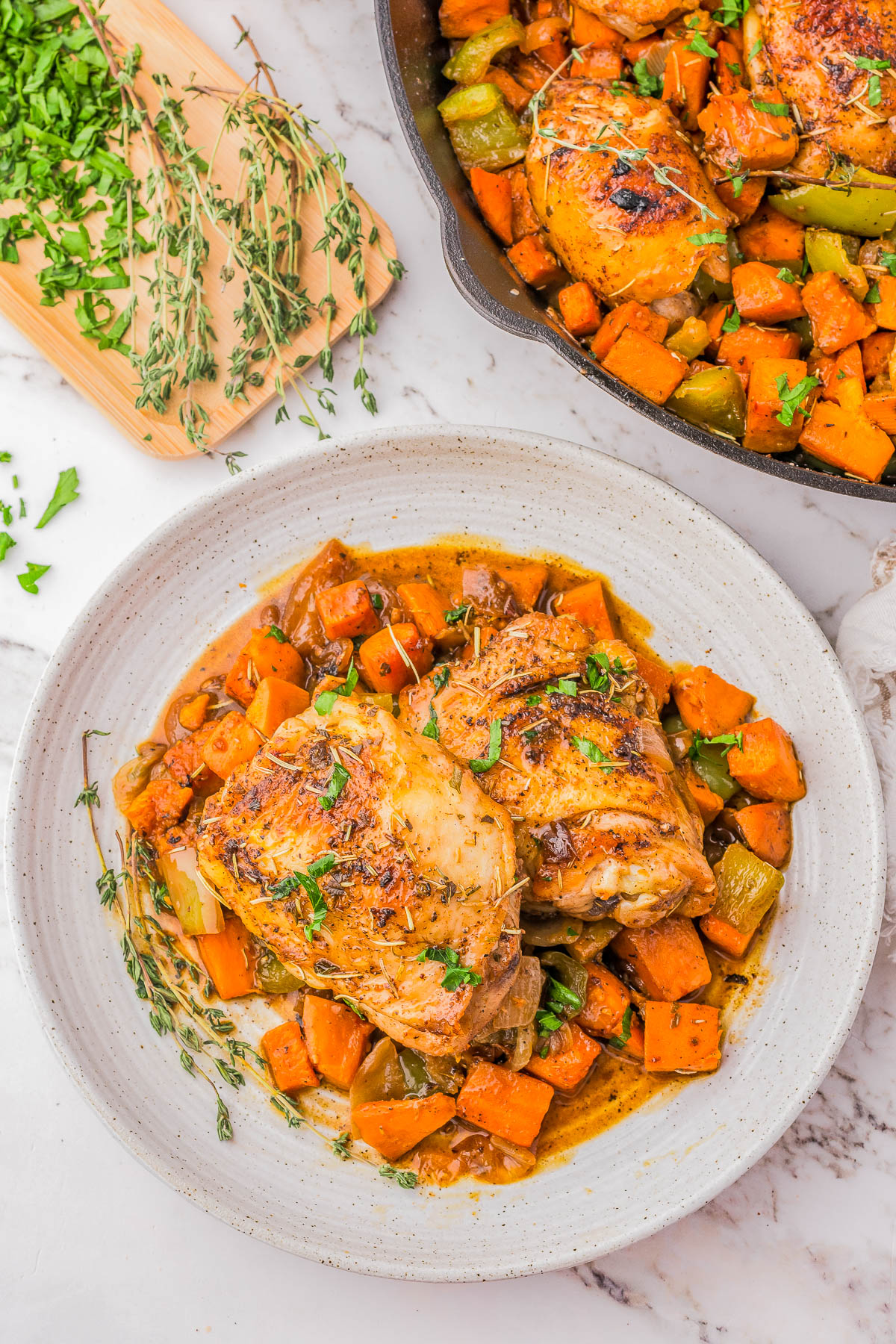 A plate of cooked chicken thighs served with diced sweet potatoes and vegetables, garnished with fresh herbs. A cutting board with green herbs and a skillet with more of the dish are in the background.