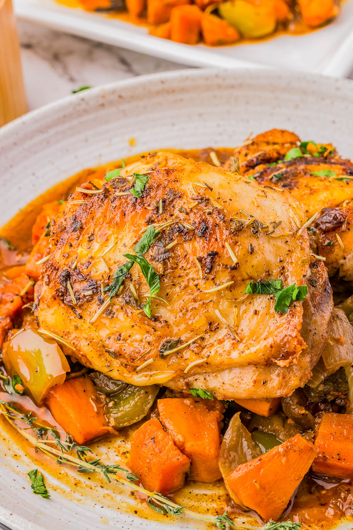 A close-up of seasoned roasted chicken thigh atop a medley of roasted vegetables, including carrots and bell peppers, garnished with fresh herbs.