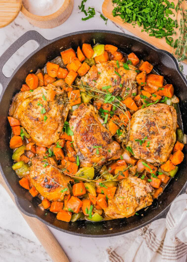 A cast-iron skillet filled with cooked chicken thighs, diced sweet potatoes, and green bell peppers, garnished with chopped parsley. A cutting board with chopped parsley and a bowl of salt are beside the skillet.