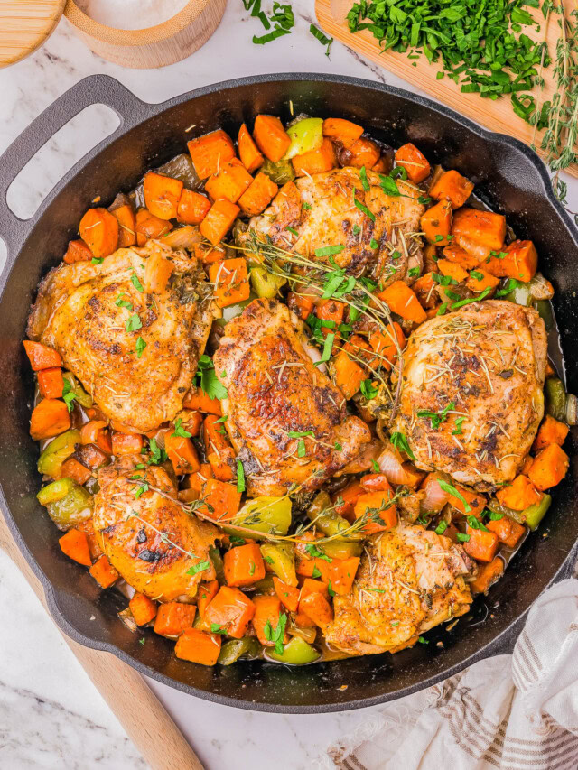 A cast-iron skillet filled with cooked chicken thighs, diced sweet potatoes, and green bell peppers, garnished with chopped parsley. A cutting board with chopped parsley and a bowl of salt are beside the skillet.