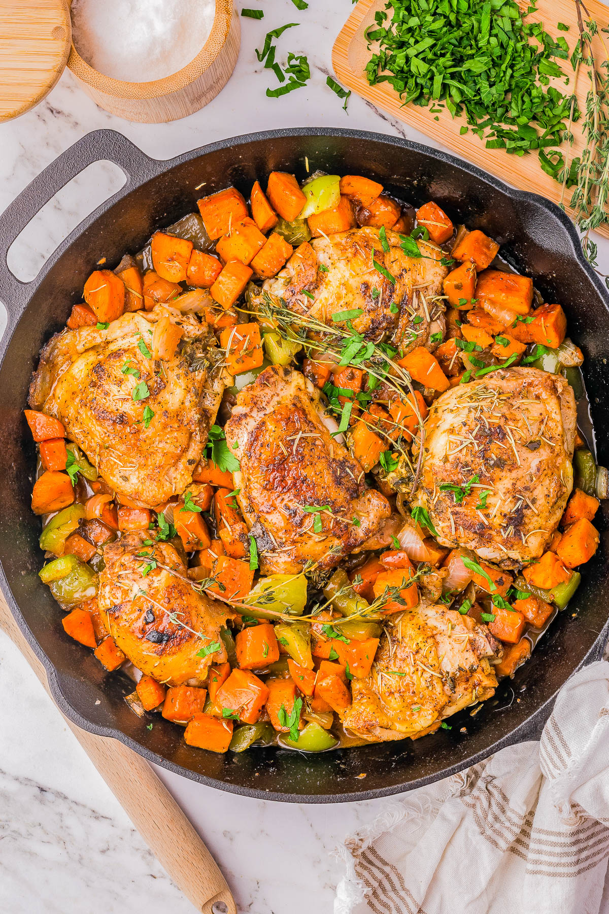 A cast-iron skillet filled with cooked chicken thighs, diced sweet potatoes, and green bell peppers, garnished with chopped parsley. A cutting board with chopped parsley and a bowl of salt are beside the skillet.