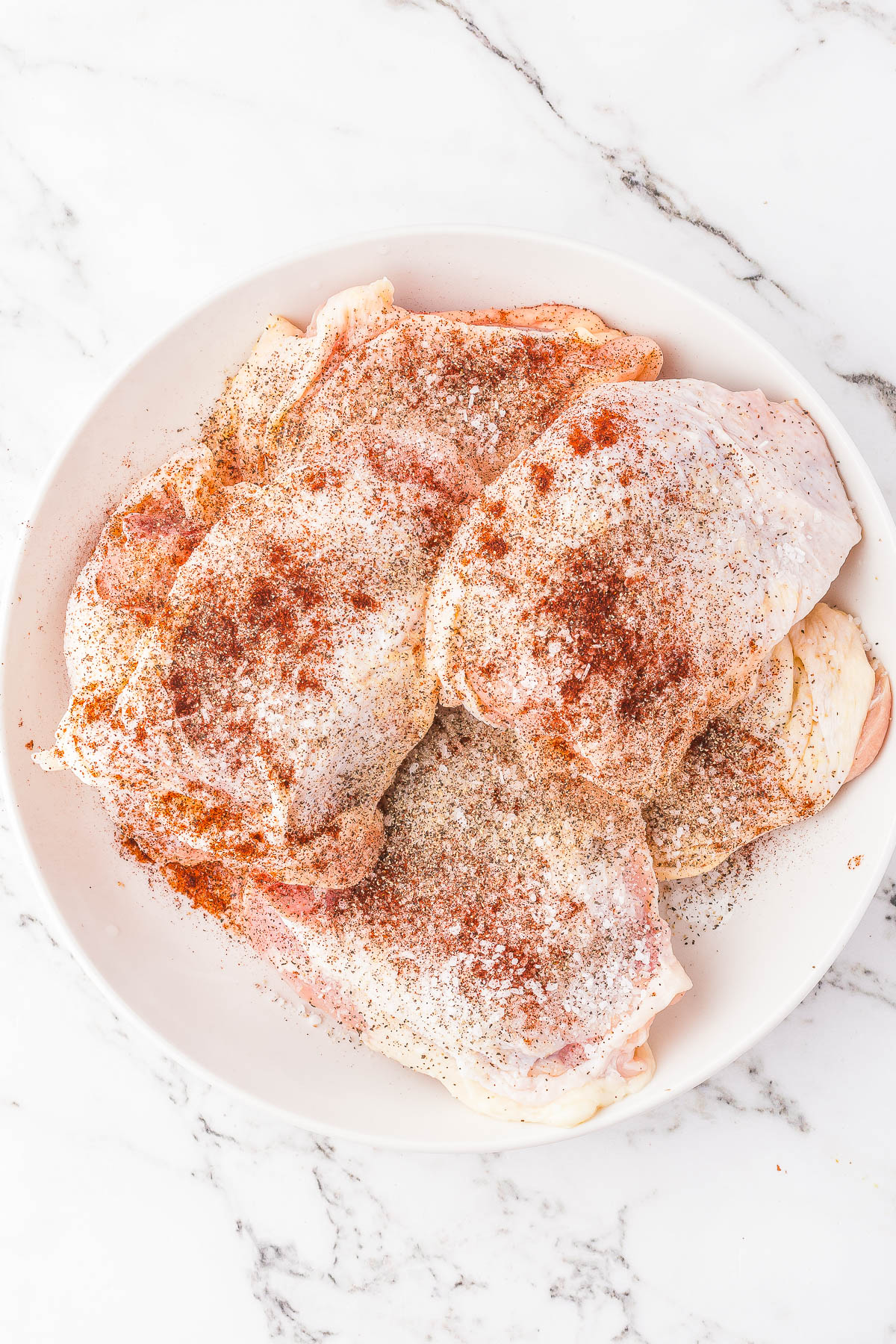 A white plate with raw, seasoned chicken thighs on a white, marble surface. The chicken is covered in various spices.