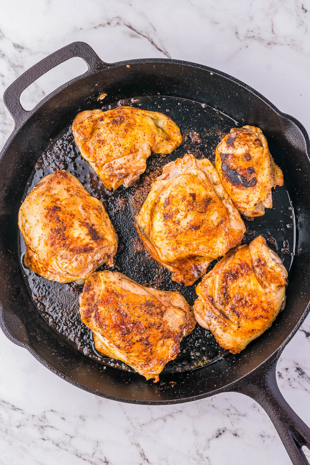 A cast iron skillet containing six pieces of browned, cooked chicken on a marble countertop.