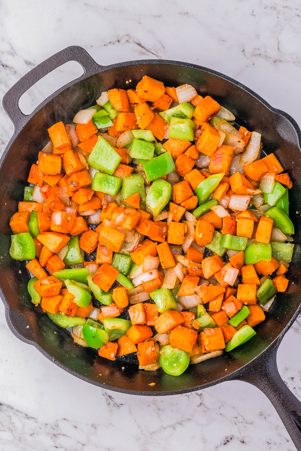 A cast iron skillet filled with diced sweet potatoes, bell peppers, and onions, sautéed and seasoned, on a marble countertop.