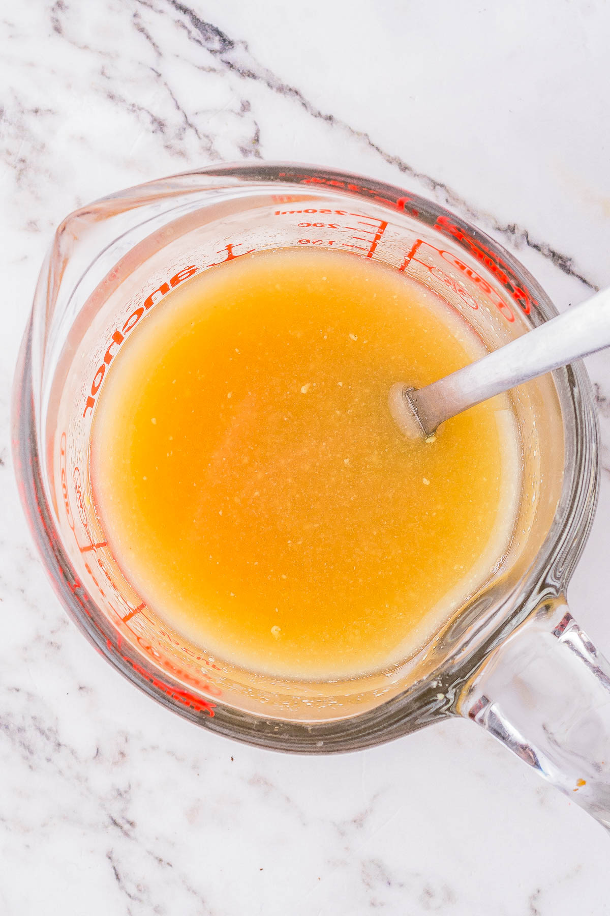 A glass measuring cup containing a light brown liquid mixture is placed on a marble surface, with a metal spoon inside.