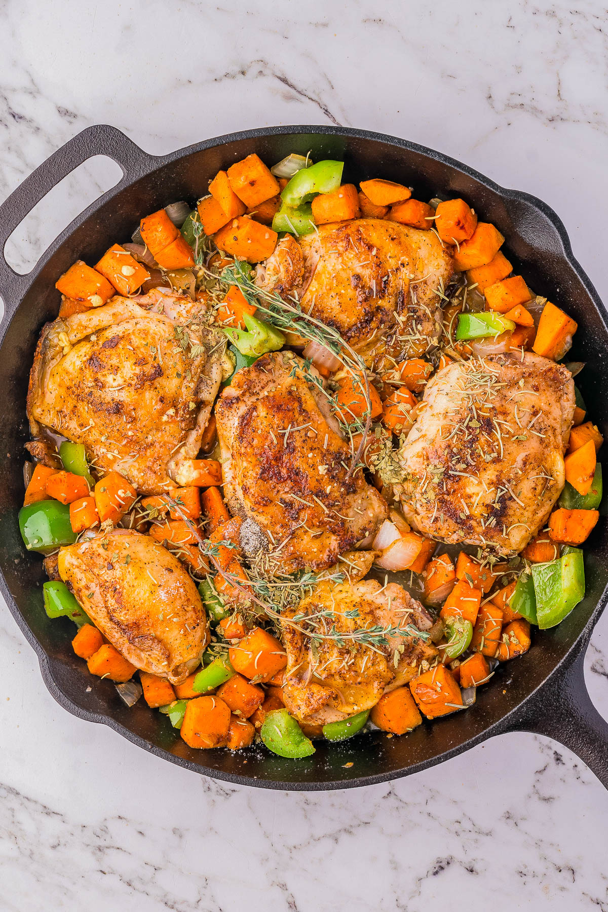 A cast iron skillet filled with seasoned, browned chicken thighs, sweet potatoes, green bell peppers, and onions, garnished with fresh herbs, placed on a marble countertop.