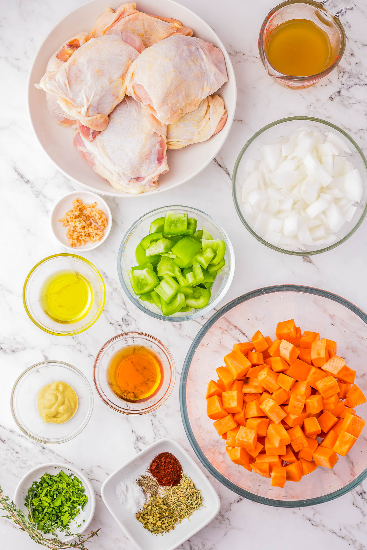 A variety of ingredients including raw chicken thighs, chopped onions, green bell peppers, diced sweet potatoes, minced garlic, olive oil, mustard, and a mix of spices are laid out on a marble surface.