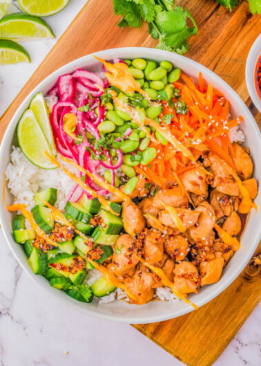 A bowl of white rice topped with cooked cubed chicken, sliced cucumbers, shredded carrots, edamame, pickled onions, lime wedges, sesame seeds, and a drizzle of sauce, garnished with cilantro.