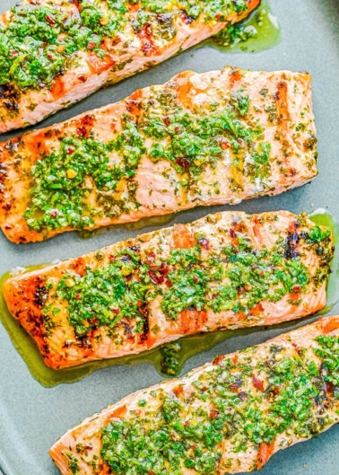 Four grilled salmon fillets topped with a green herb sauce on a gray plate.