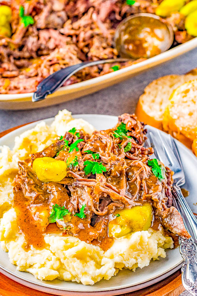 A plate of mashed potatoes topped with shredded beef and garnished with parsley. A serving dish with more beef and a ladle is visible in the background. A slice of bread is on the side.