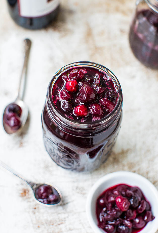 A glass jar filled with dark berry preserve, surrounded by a small white bowl and a spoon containing some of the preserve, set on a rustic white surface.