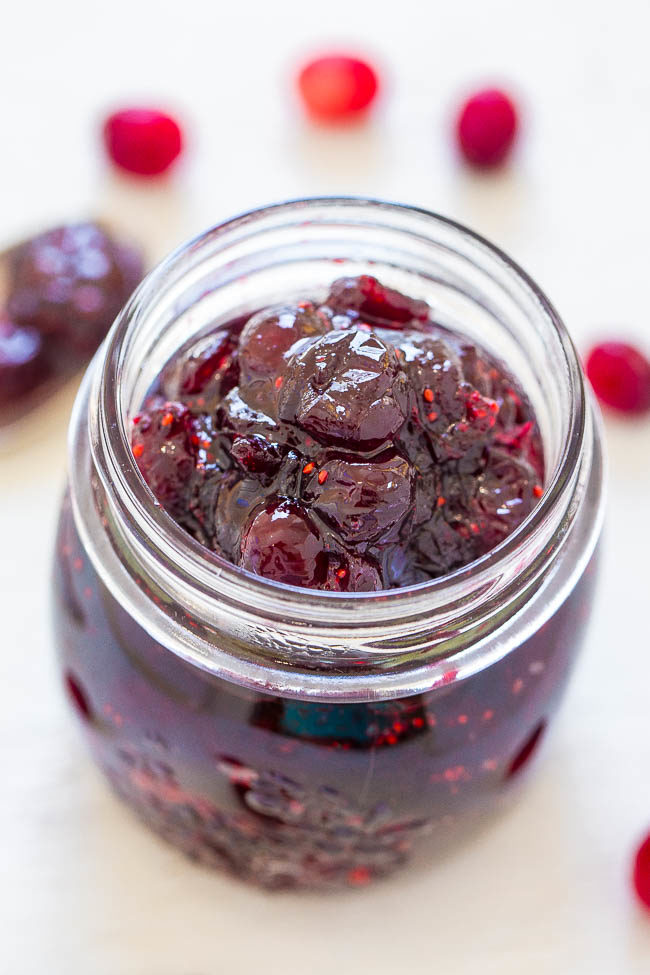 A glass jar filled with dark red cranberry jam, with a few whole cranberries scattered around it.