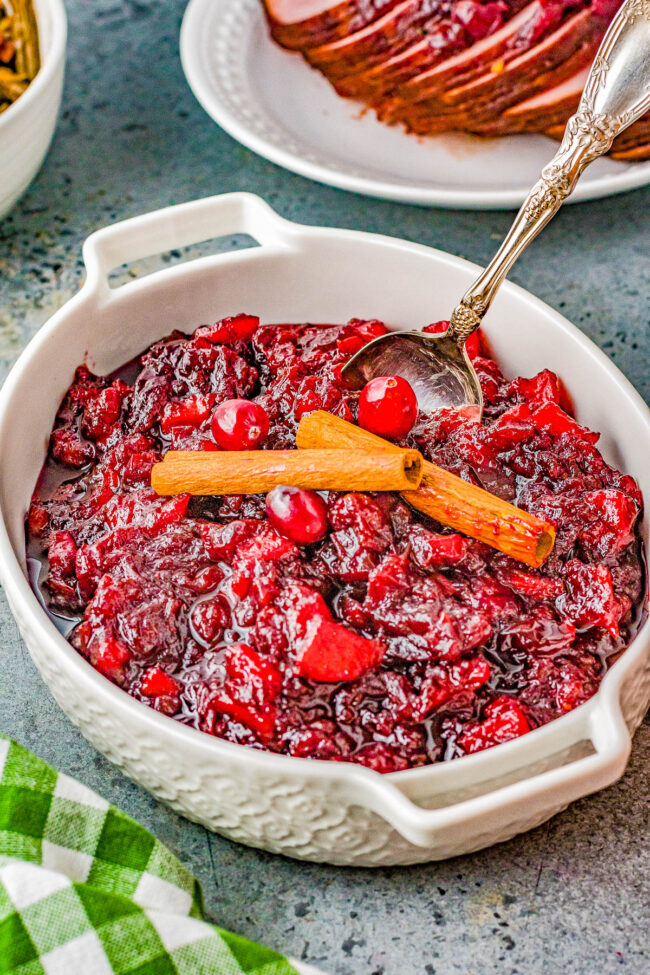 A white dish filled with cranberry sauce garnished with whole cranberries and cinnamon sticks, with a spoon resting inside.