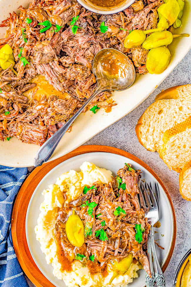 A plate of shredded beef with gravy and yellow peppers served over mashed potatoes, garnished with parsley. Next to it is a serving platter with more beef and a bread slice.