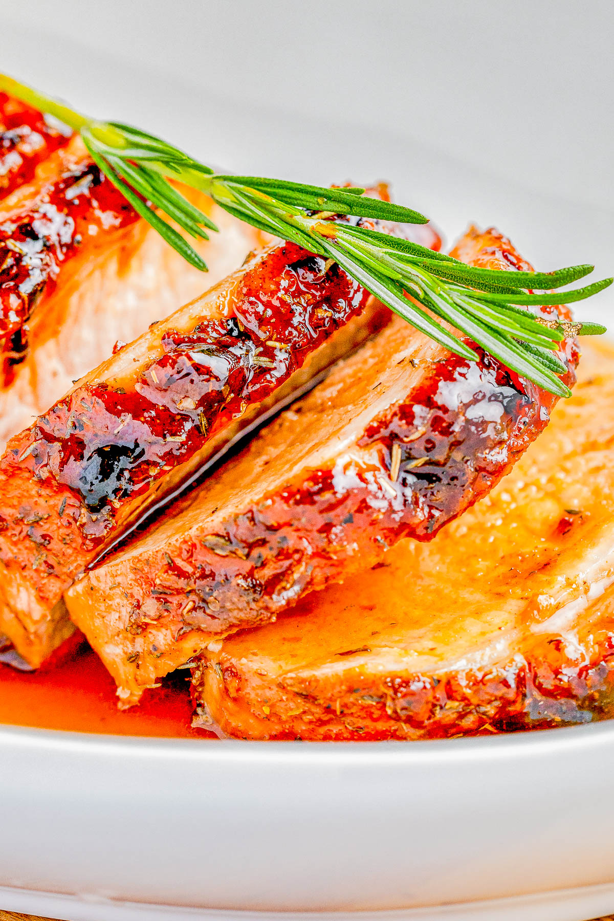 Close-up of crispy, glazed slices of roasted meat garnished with a sprig of rosemary, served on a white plate.