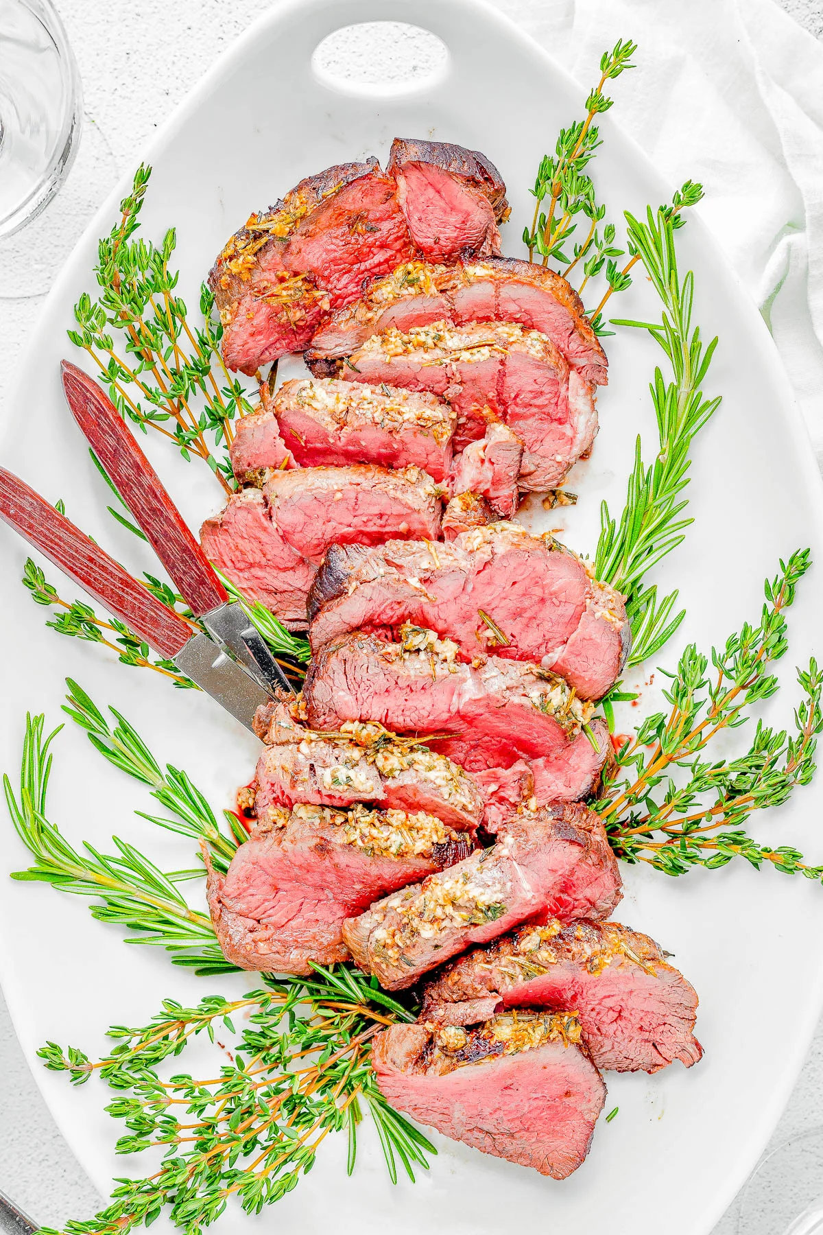 A white oval dish with several slices of medium-rare beef tenderloin garnished with fresh rosemary sprigs. A carving fork is resting on the dish.