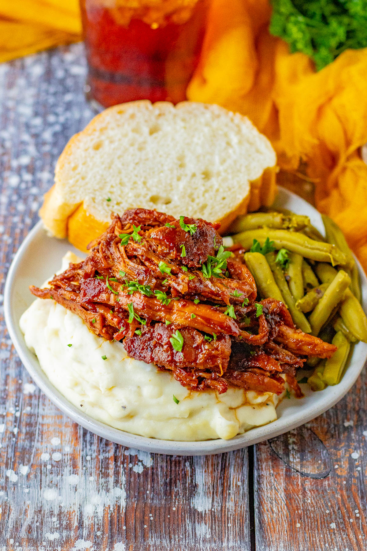 A plate of shredded barbecue meat sits atop mashed potatoes with a side of green beans, garnished with chopped herbs. Two slices of bread are placed behind the meal, and a drink is partially visible.