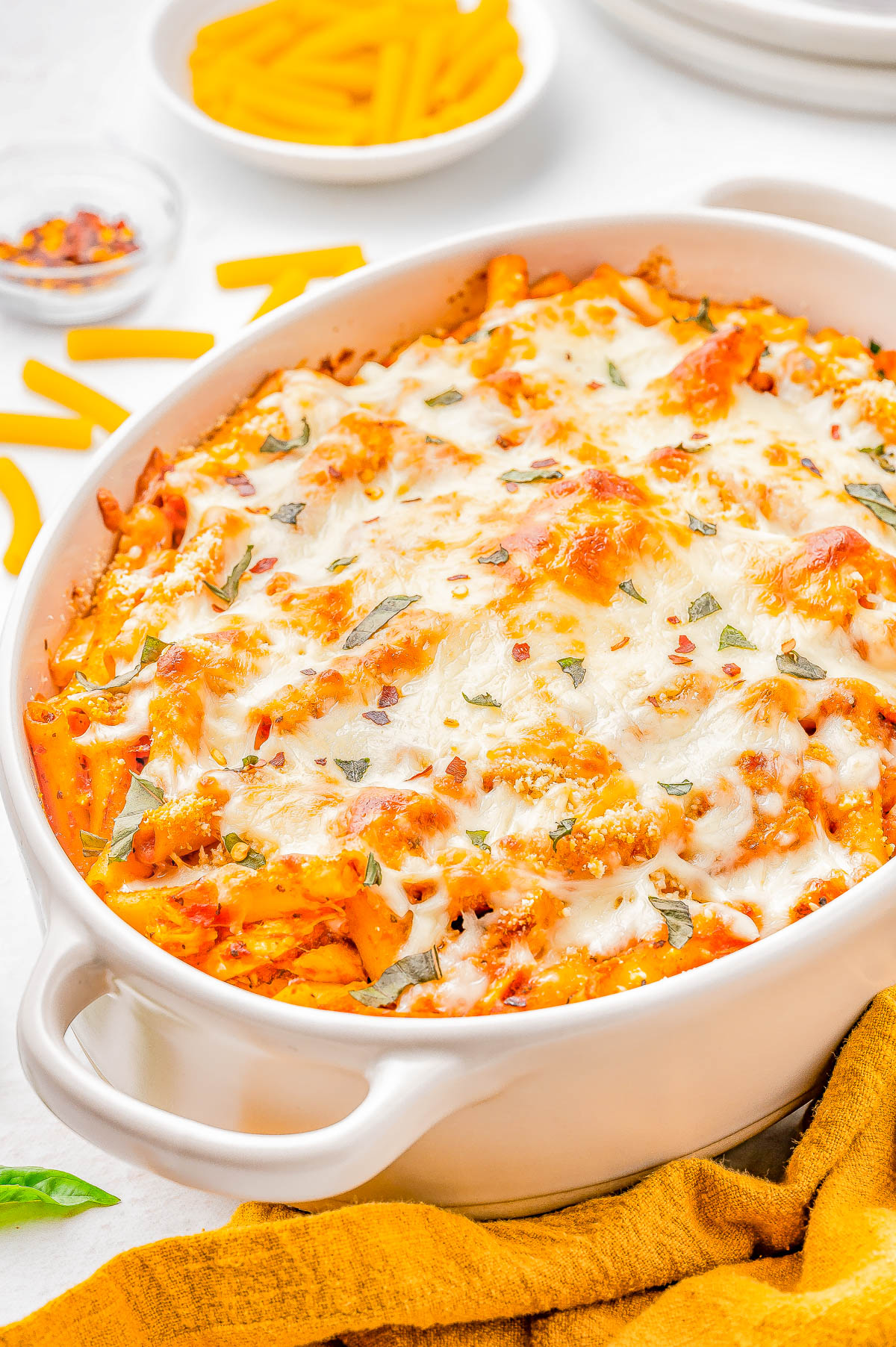 Baked pasta with melted cheese and herbs in a white dish, surrounded by a yellow napkin, uncooked pasta, and a small dish of red pepper flakes.