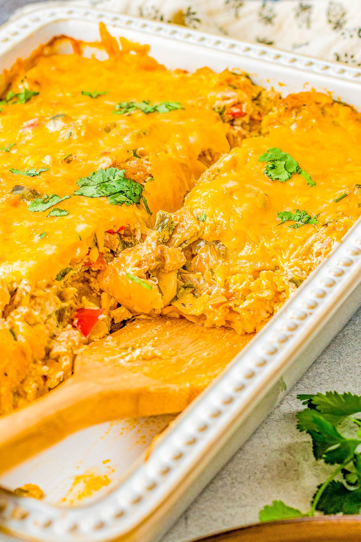 A casserole dish with a cheesy baked casserole topped with cilantro. A portion is cut out, revealing layers of cheese, rice, and vegetables. A wooden spatula rests in the dish.