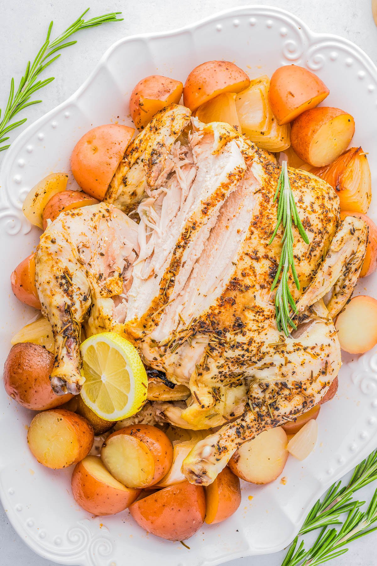 Roasted chicken on a white platter surrounded by halved potatoes, garnished with rosemary sprigs and a lemon slice.