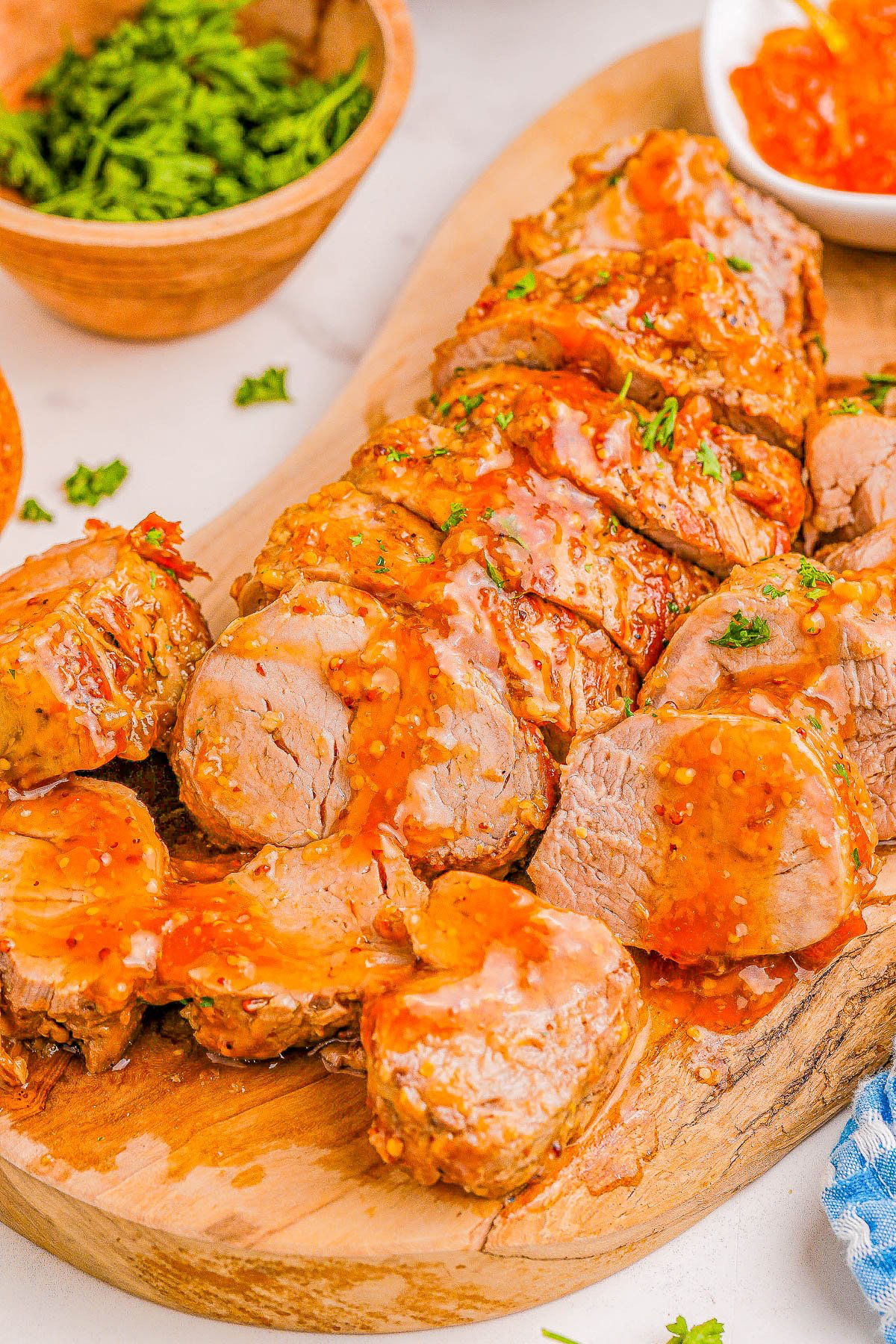 Sliced pork tenderloin with a glazed sauce on a wooden board, garnished with parsley, with a bowl of greens and a sauce dish in the background.