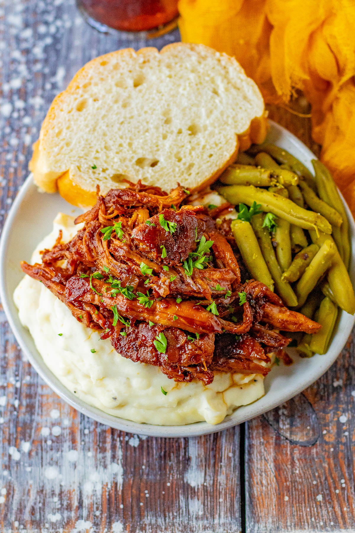 A plate with barbecued pulled pork over mashed potatoes, green beans, and a slice of bread, garnished with chopped parsley.