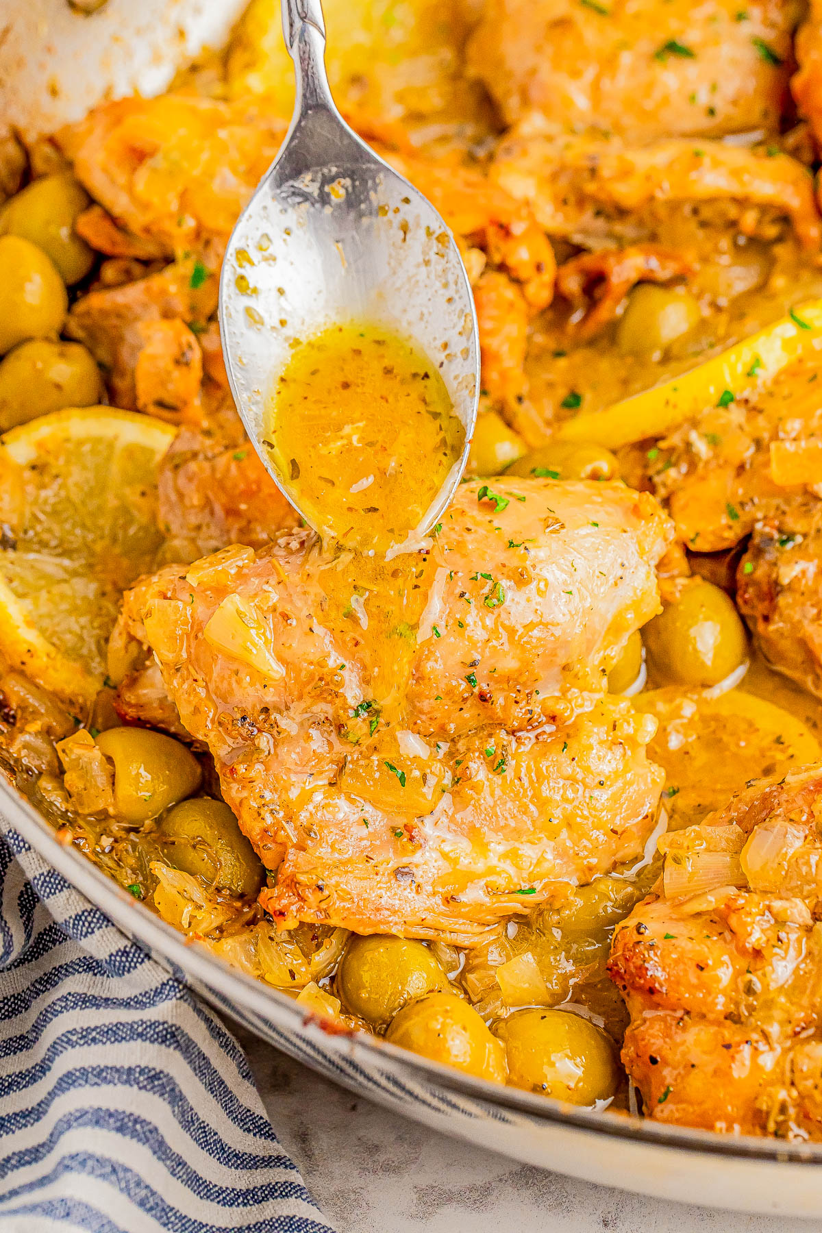 A spoon drizzles sauce over cooked chicken thighs with green olives and lemon slices in a pan.