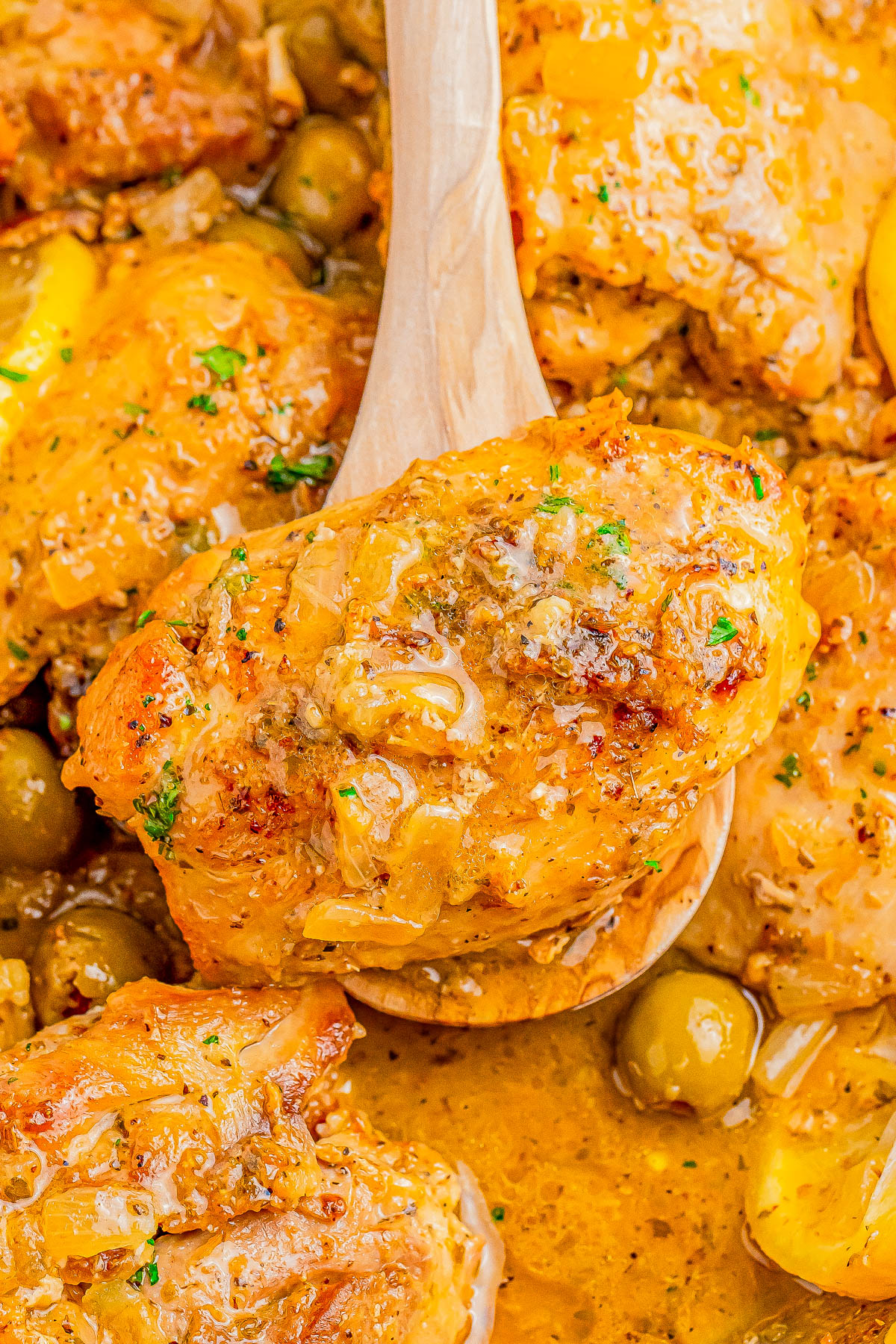 Close-up of a wooden spoon lifting a piece of seasoned and cooked chicken from a dish with olives and sauce.