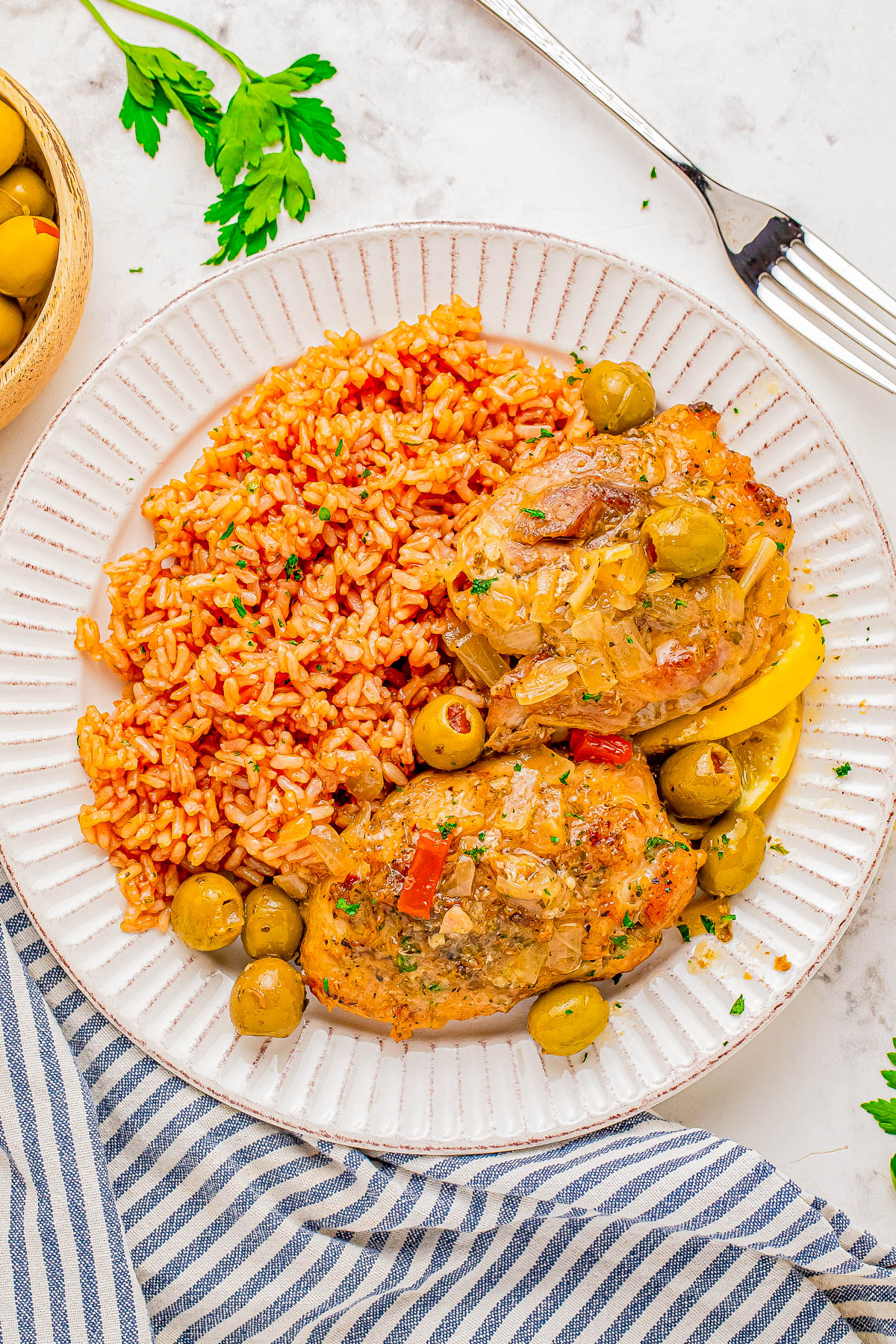 A plate with seasoned rice, two pieces of chicken topped with a sauce and olives, garnished with parsley, next to a fork. A blue-striped napkin and a bowl of olives are in the background.