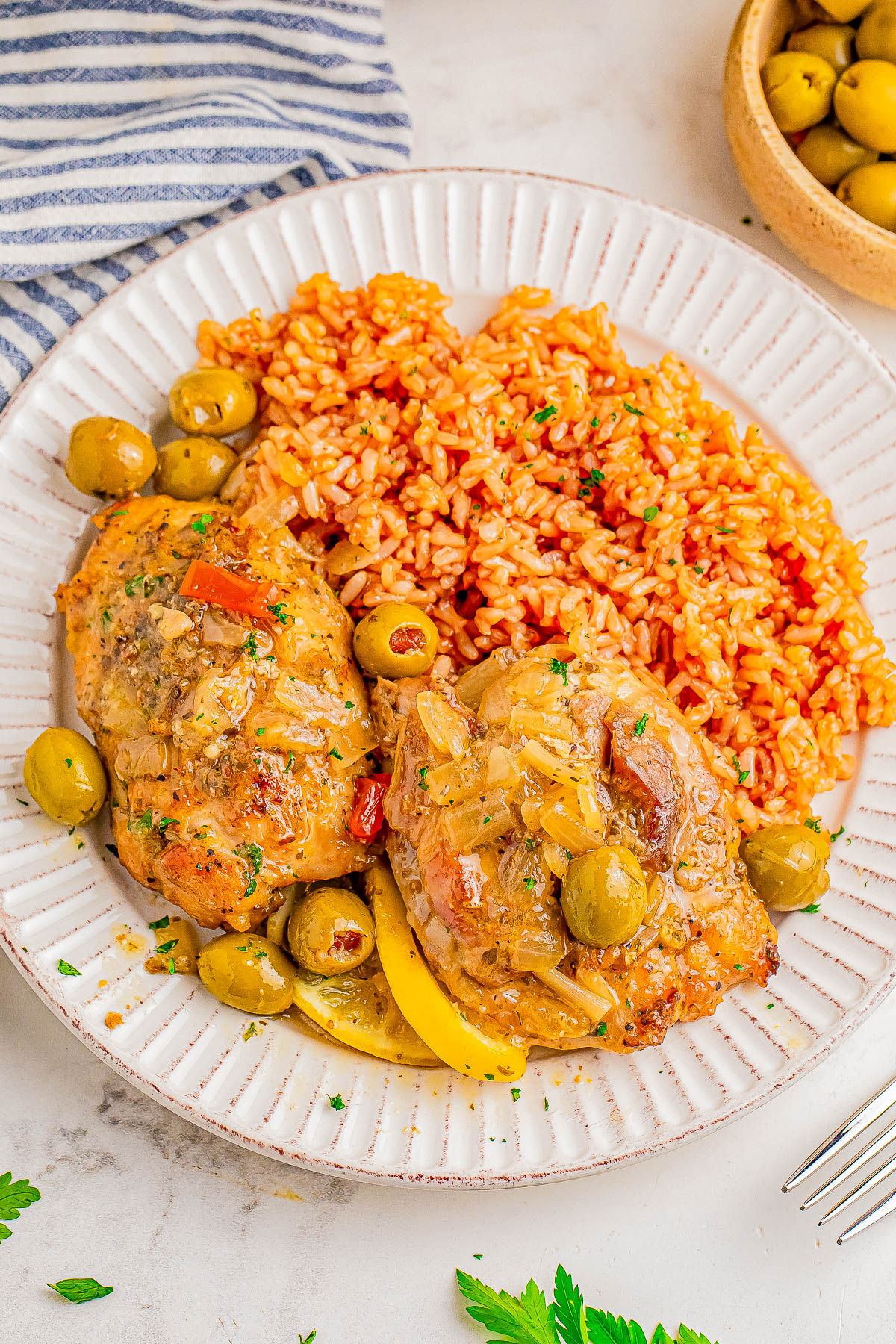 A plate with two pieces of chicken covered in a sauce with olives, and a side of seasoned rice. A bowl of green olives and a striped napkin are beside the plate.