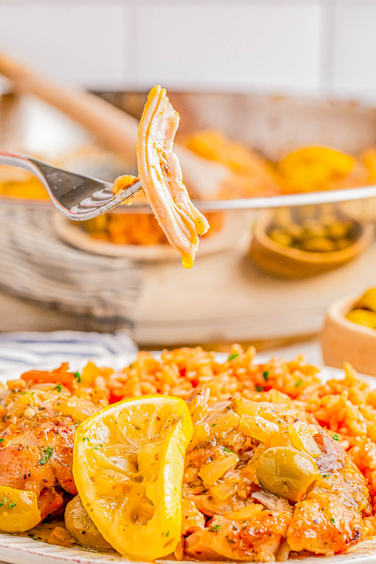 A fork with a piece of chicken is held above a plate of cooked chicken, rice, and lemon slices. In the background, a pan and bowls with additional ingredients are visible.