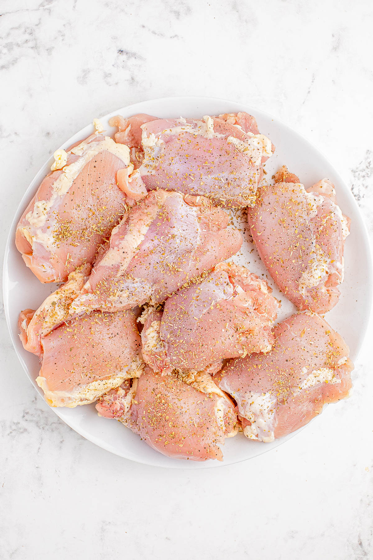 A white plate with eight raw chicken thighs seasoned with spices, set on a white marble surface.