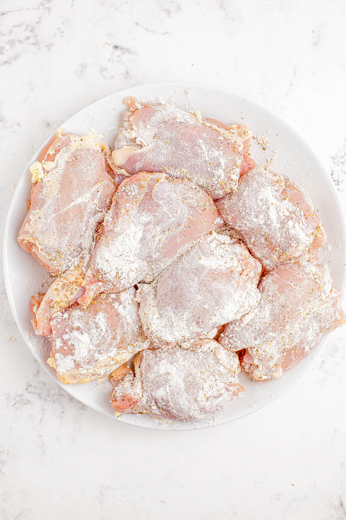 A white plate containing several raw, flour-coated chicken pieces on a marble surface.