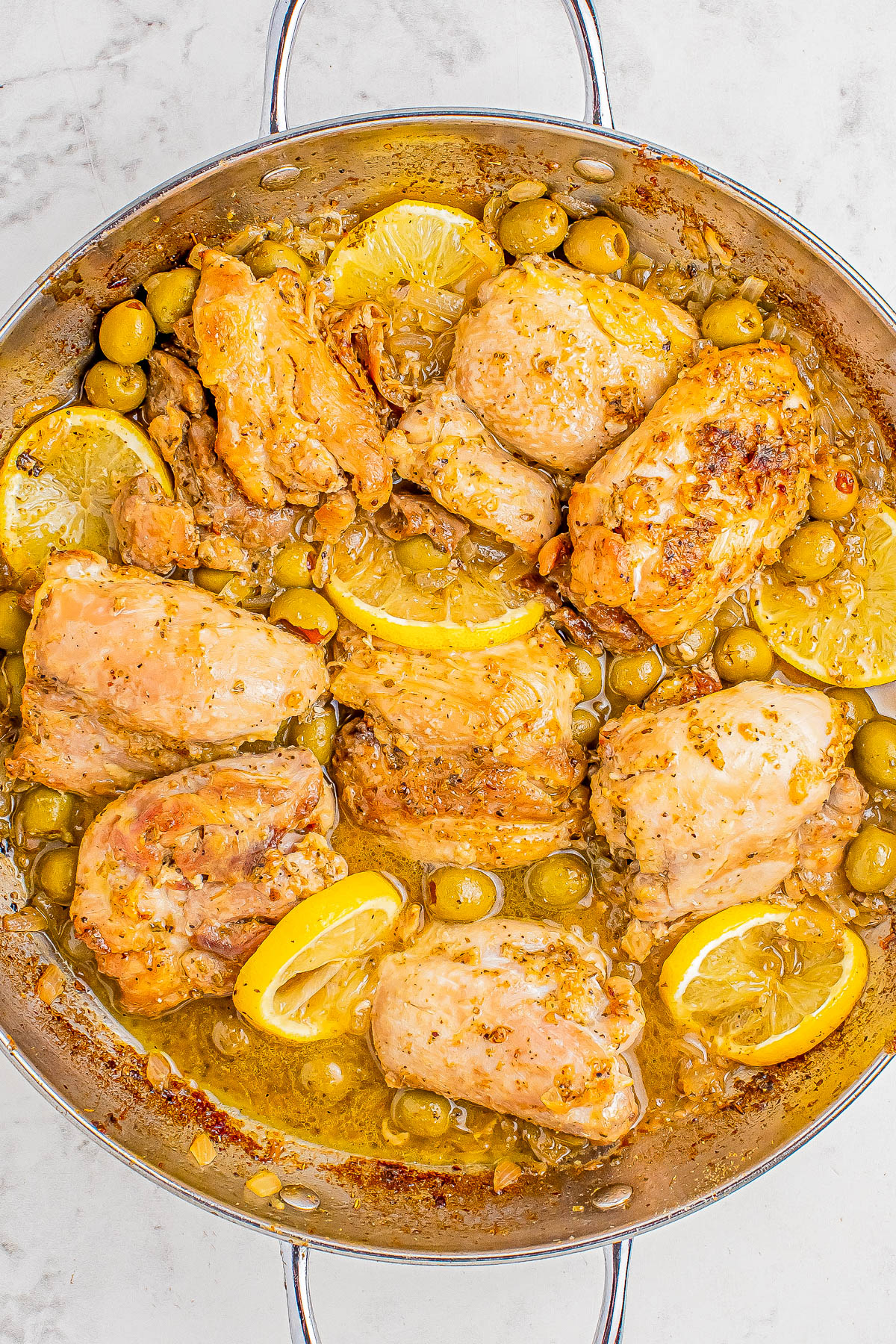 A large skillet filled with cooked chicken thighs, lemon slices, and green olives in a sauce.