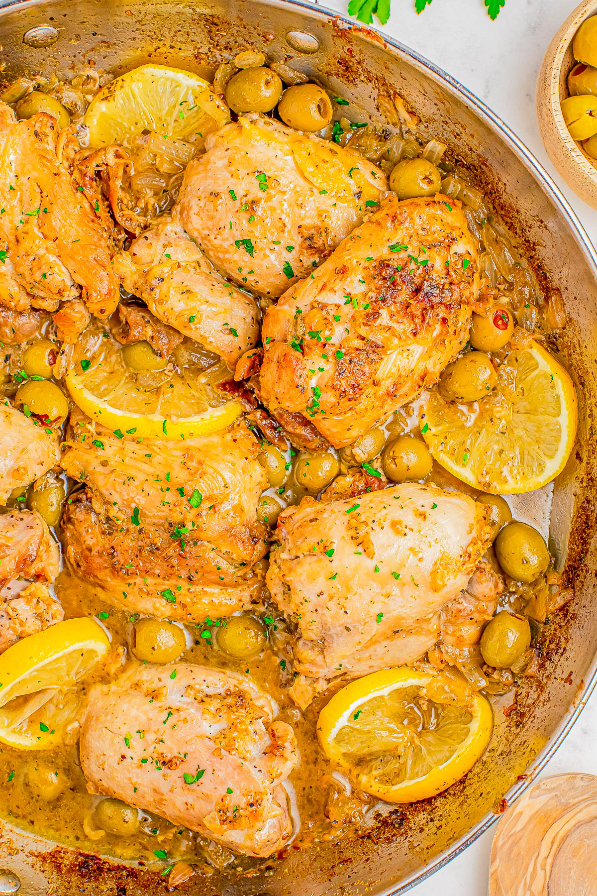 A skillet filled with cooked chicken thighs, lemon slices, green olives, and a sauce, garnished with chopped parsley.