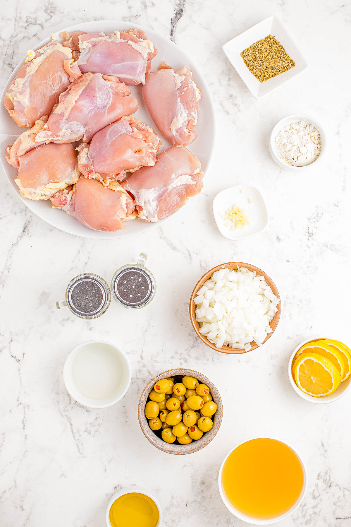 Top-down view of various cooking ingredients including raw chicken thighs, chopped onions, green olives, lemon slices, spices, and liquids arranged on a white marble surface.