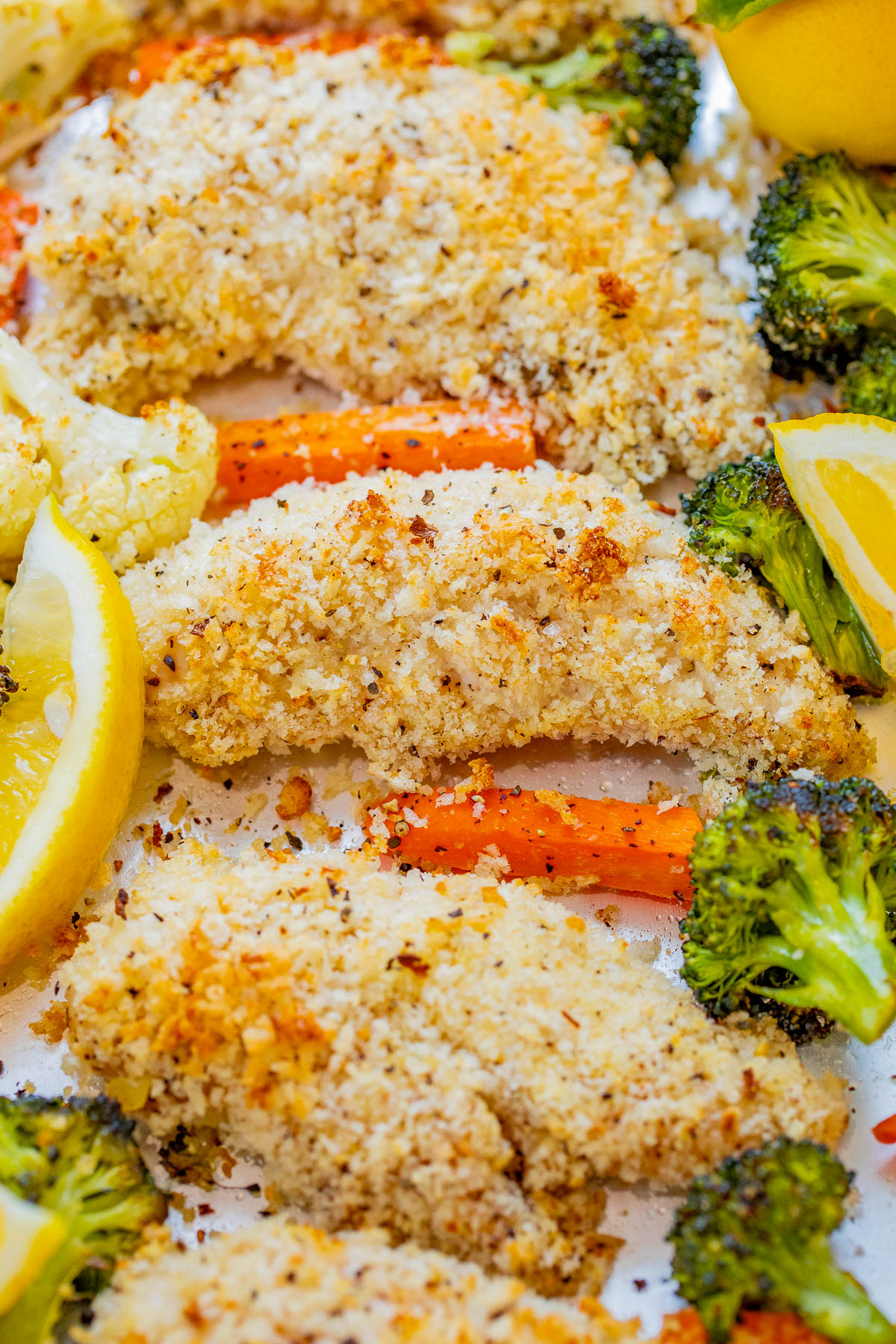 Breaded chicken tenders arranged on a baking sheet with roasted broccoli, cauliflower, carrots, and lemon wedges.