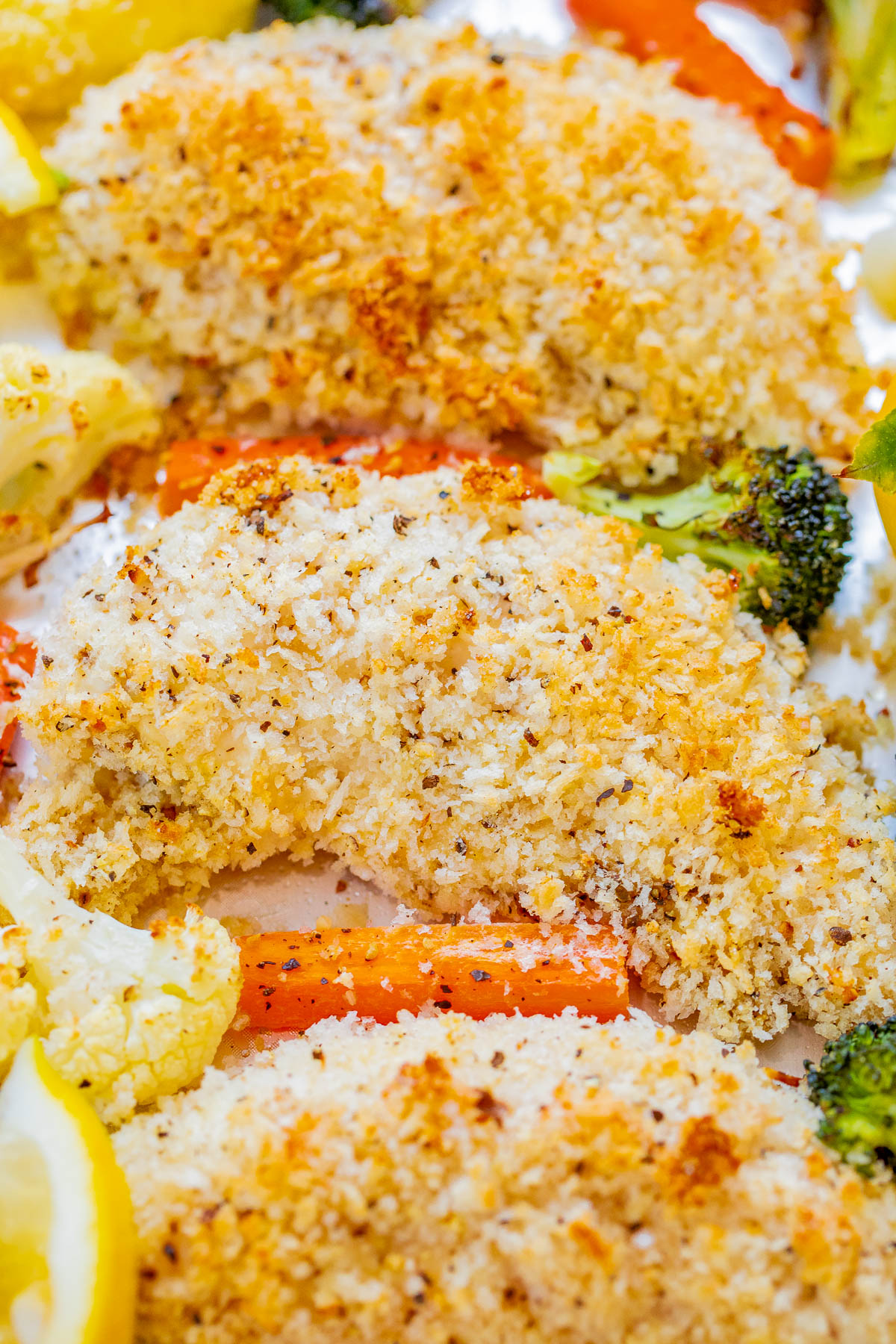 Close-up of breaded and baked chicken fillets served with roasted vegetables, including broccoli, carrots, and cauliflower, garnished with lemon slices.