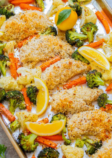 Sheet pan meal of lemon-baked chicken with breaded coating, surrounded by roasted vegetables including broccoli, carrots, and cauliflower, garnished with lemon wedges.