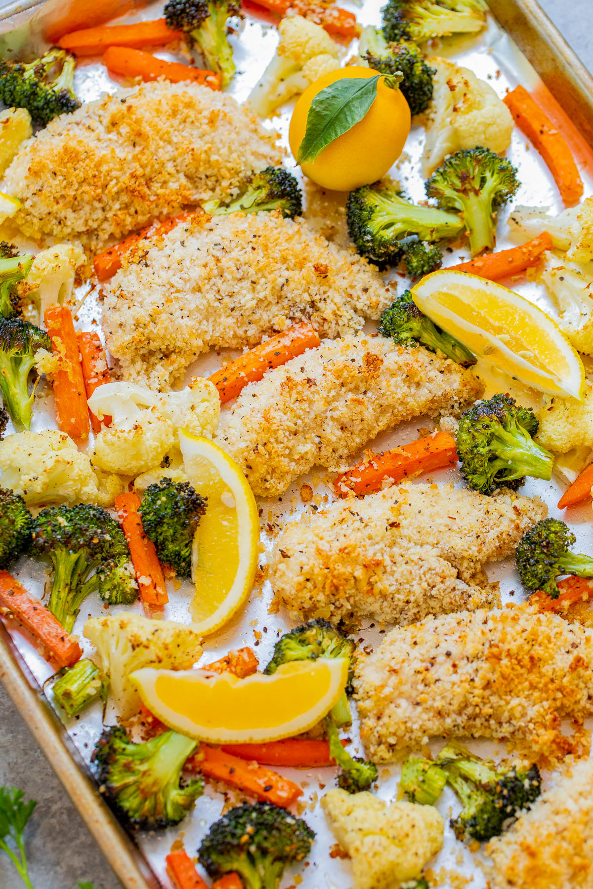 Sheet pan meal of lemon-baked chicken with breaded coating, surrounded by roasted vegetables including broccoli, carrots, and cauliflower, garnished with lemon wedges.