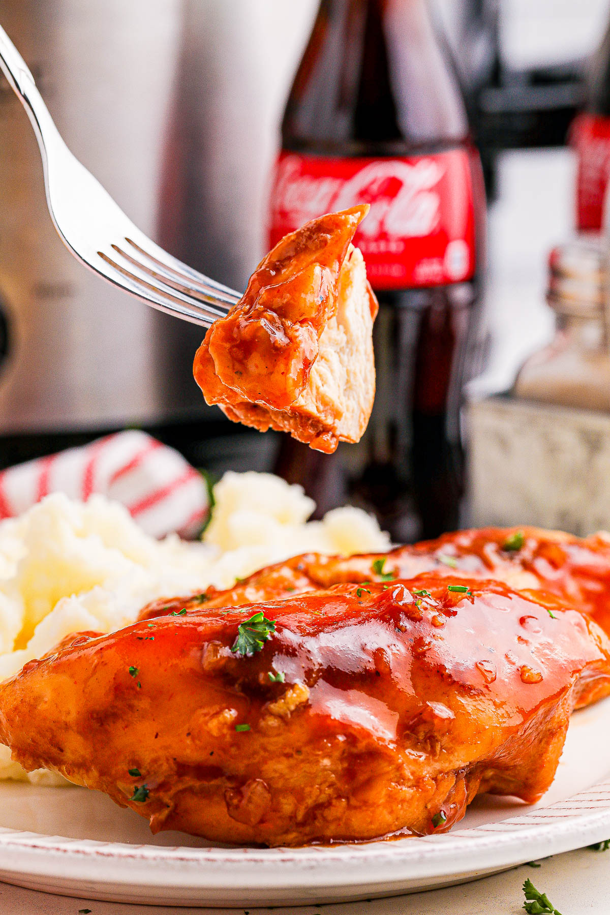 A fork holding a piece of barbecued chicken over a plate with more chicken and mashed potatoes. Two bottles of Coca-Cola are in the background.