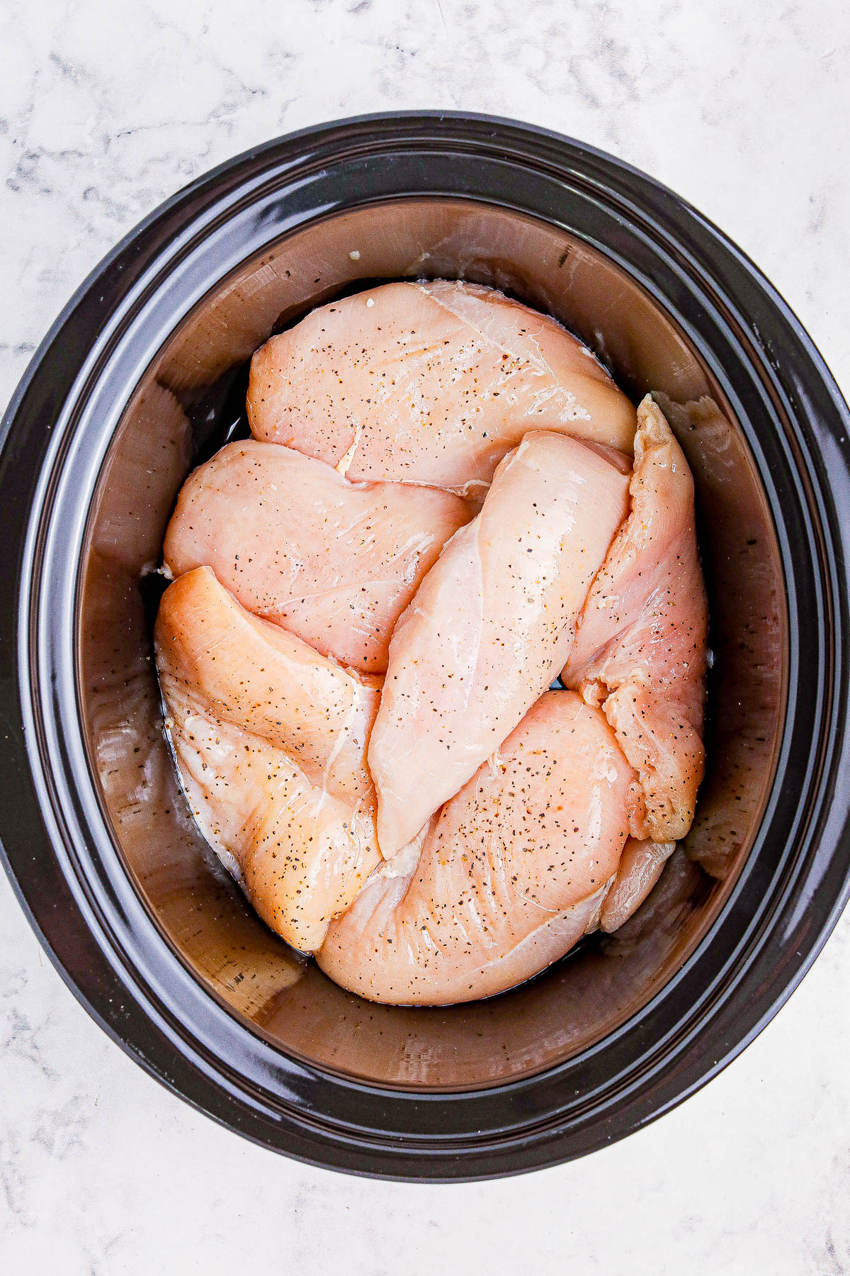 Raw chicken breasts seasoned with pepper are arranged in a black slow cooker on a marble countertop.