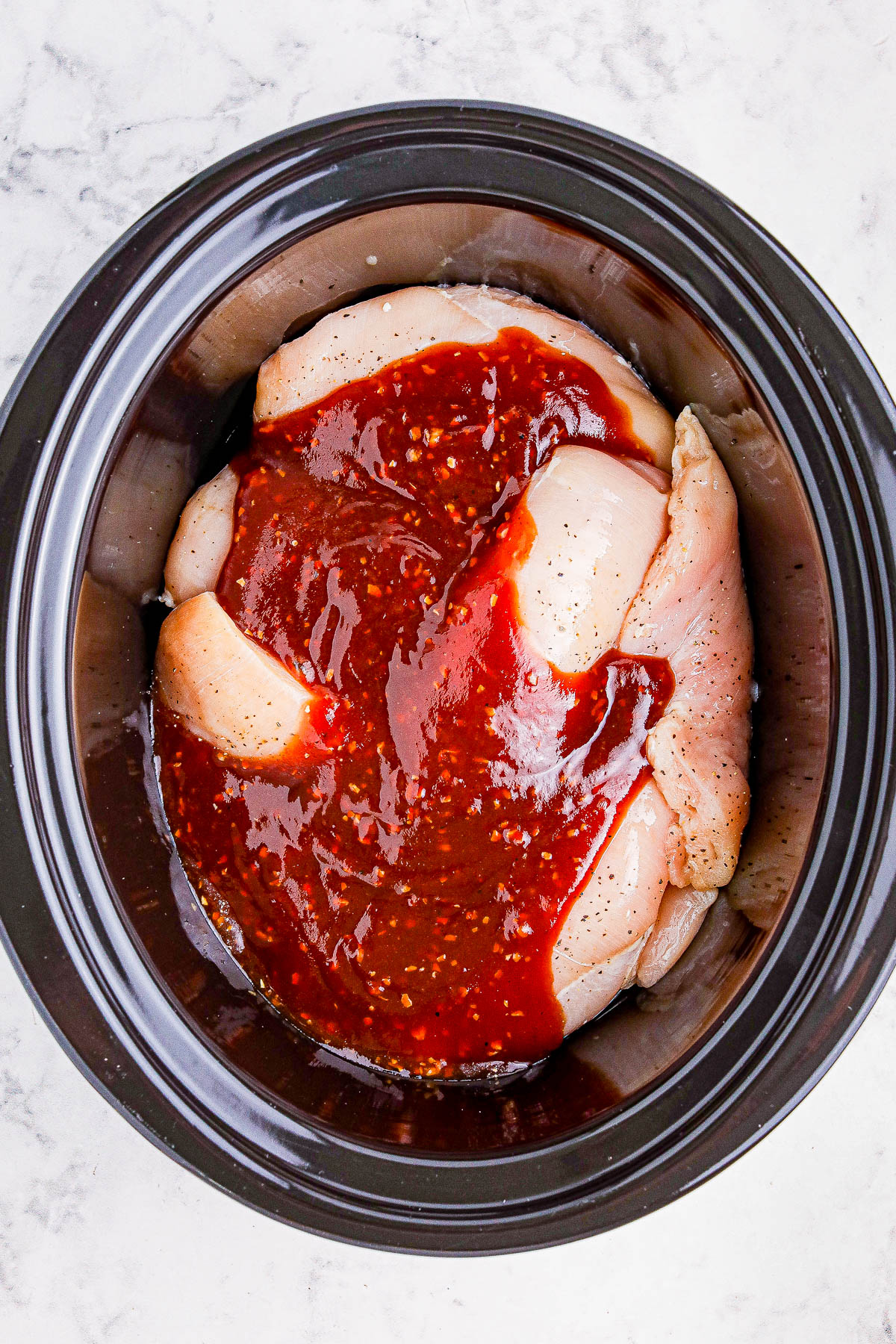 Raw chicken breasts marinated with a red sauce inside a black slow cooker, placed on a white surface.