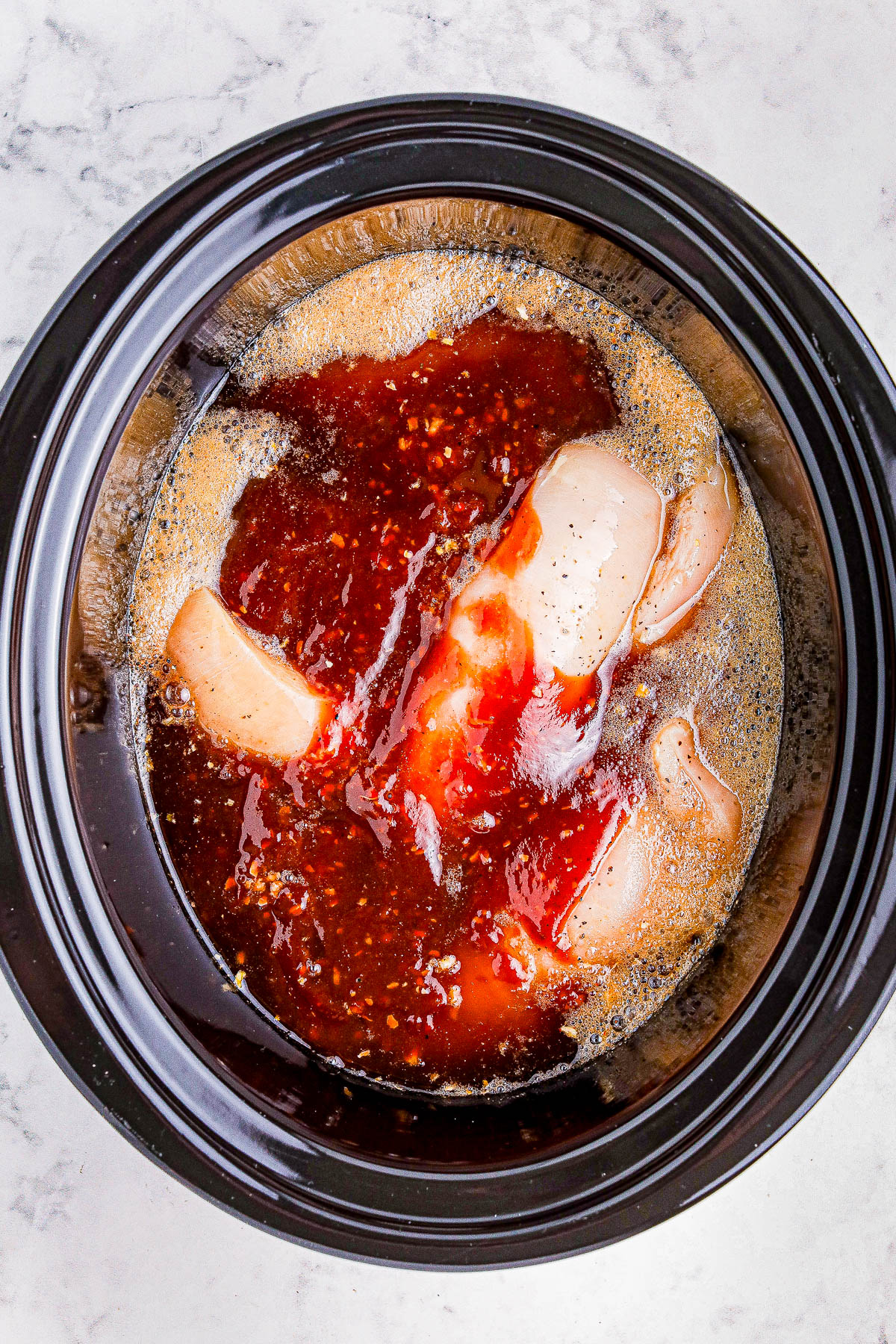 A crockpot containing raw chicken pieces submerged in a red marinade sauce, placed on a marbled countertop.