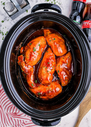 Slow cooker with five pieces of chicken breast coated in a red sauce, on a countertop with a striped towel, wooden spoon, salt and pepper shakers, and two cola bottles in the background.