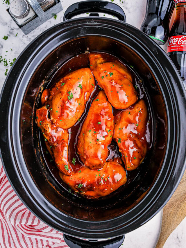 Slow cooker with five pieces of chicken breast coated in a red sauce, on a countertop with a striped towel, wooden spoon, salt and pepper shakers, and two cola bottles in the background.