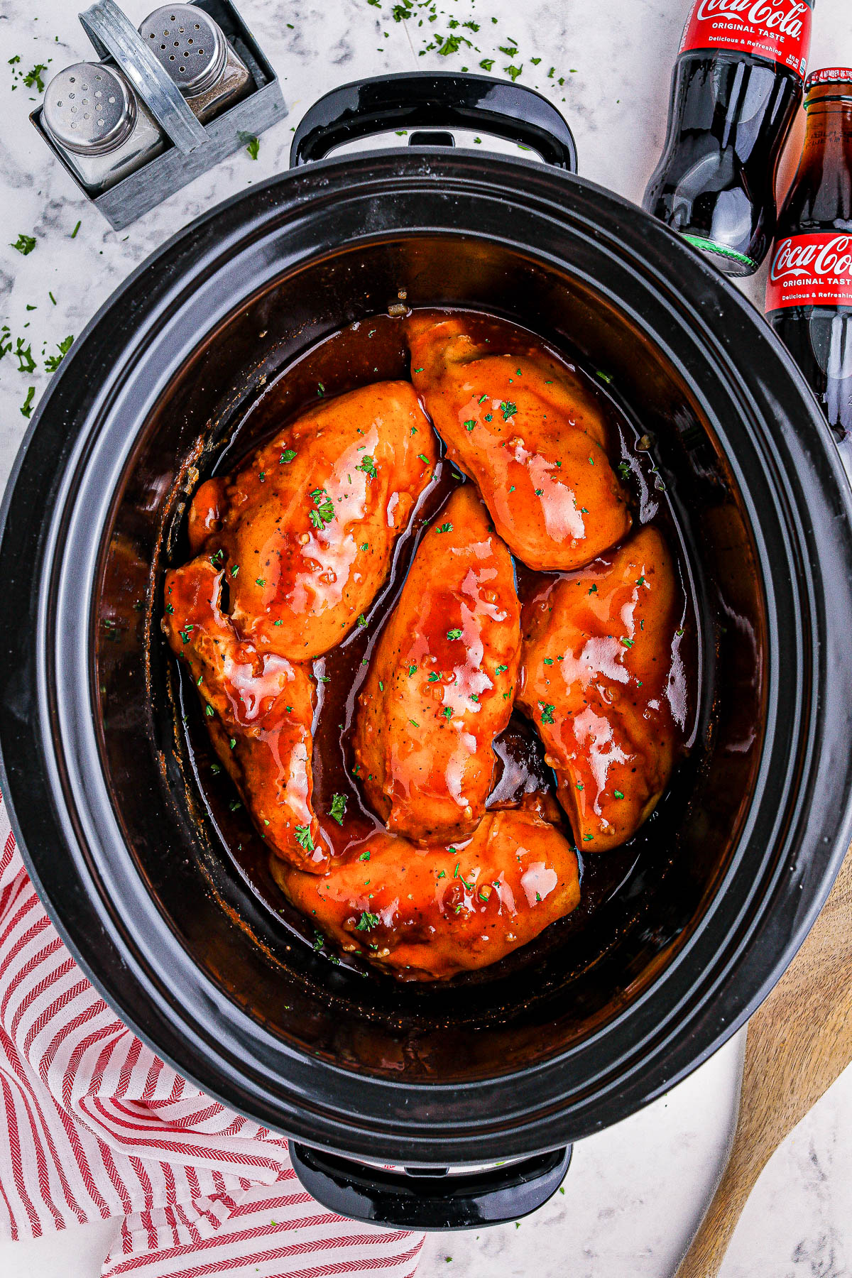 Slow cooker with five pieces of chicken breast coated in a red sauce, on a countertop with a striped towel, wooden spoon, salt and pepper shakers, and two cola bottles in the background.